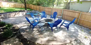 Round paver patio with a fire pit and Adirondack chairs inside a fenced backyard
